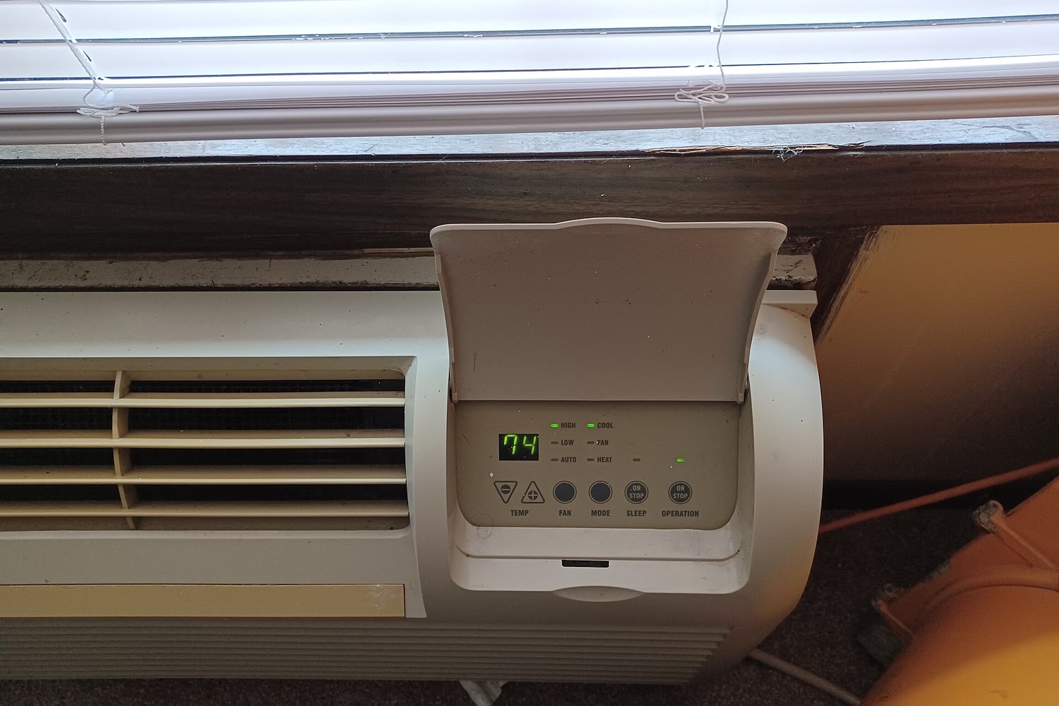 Packaged terminal air conditioner (PTAC) under a window with its control door open, showing the set temperature and mode buttons