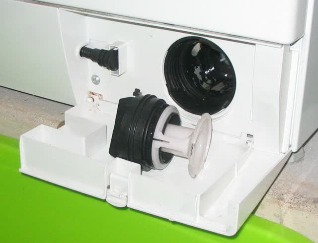 Front-load washer kick panel open: drain pump filter (coin trap) unscrewed and pulled out beside its housing, with the small emergency drain hose on the left